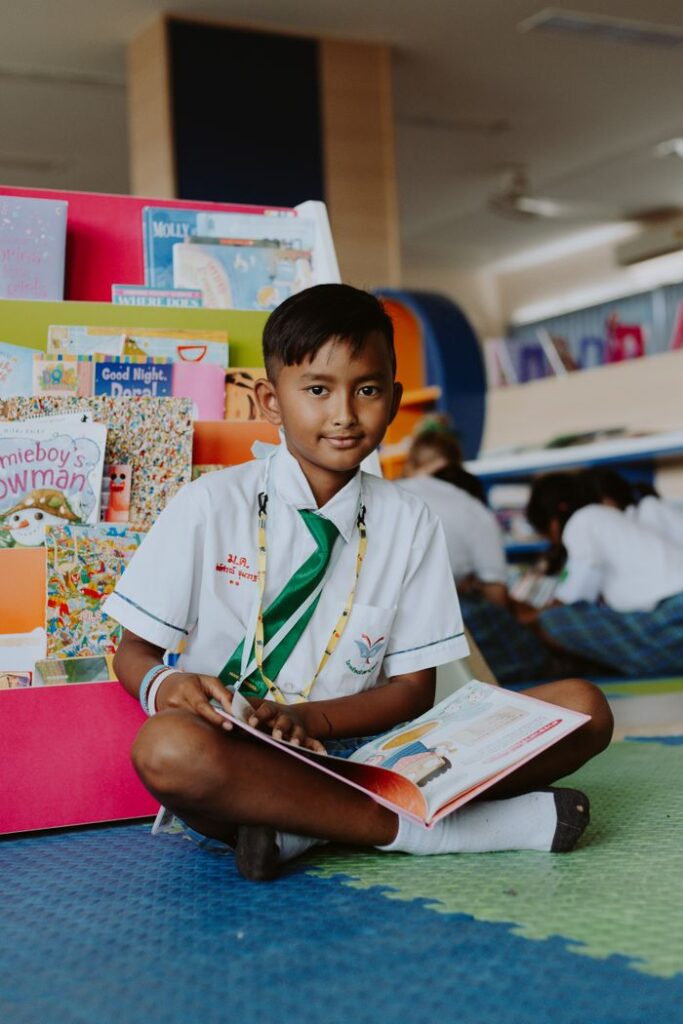 A boy in a school uniform sits cross-legged reading a book in a classroom.