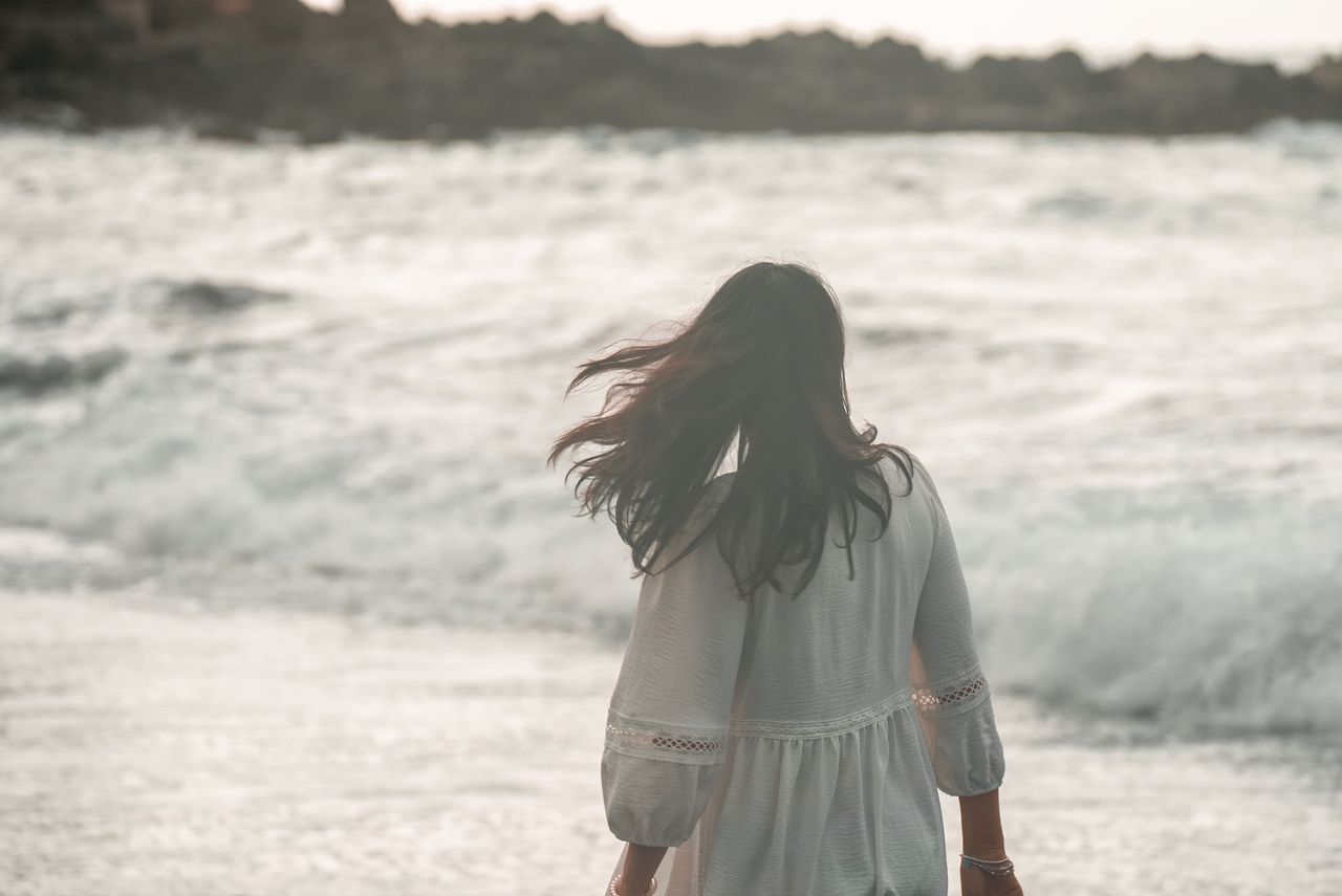 Woman in white dress walks by a stormy sea with waves crashing against the shore.