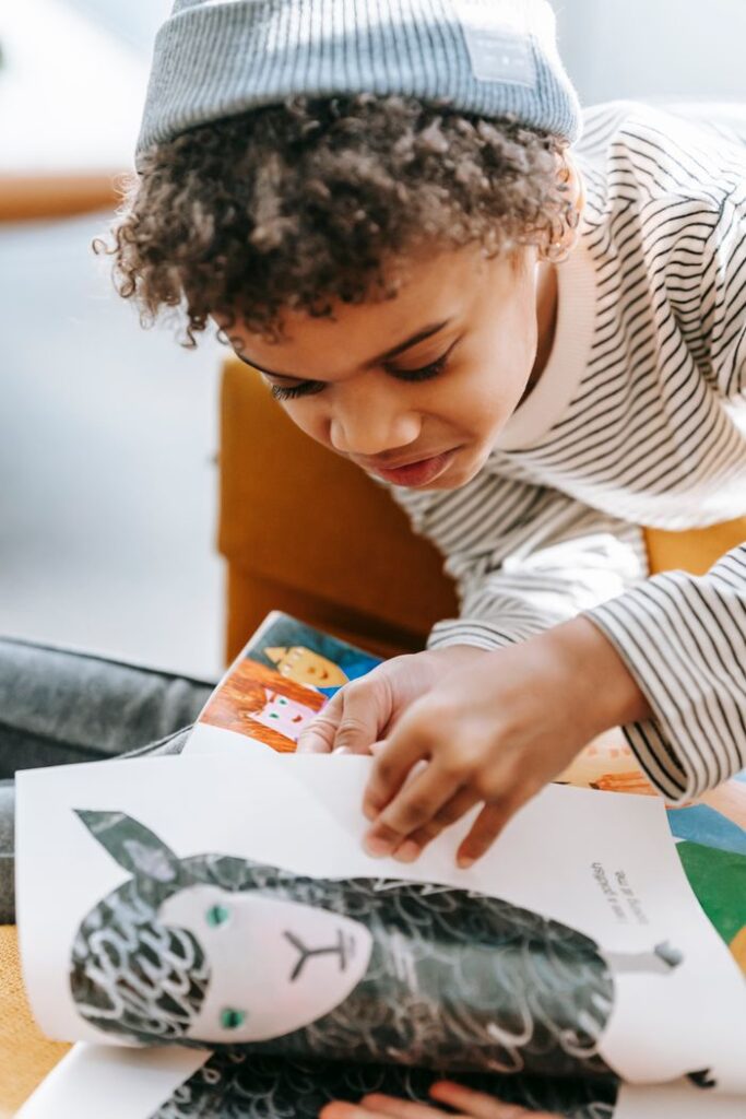 Curious African American boy wearing casual clothes and hat turning pages of book with picture of sheep in light room