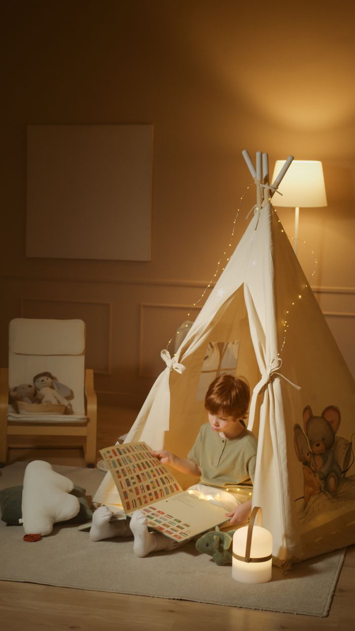 Young boy reads a book in a cozy indoor tent with warm lighting.